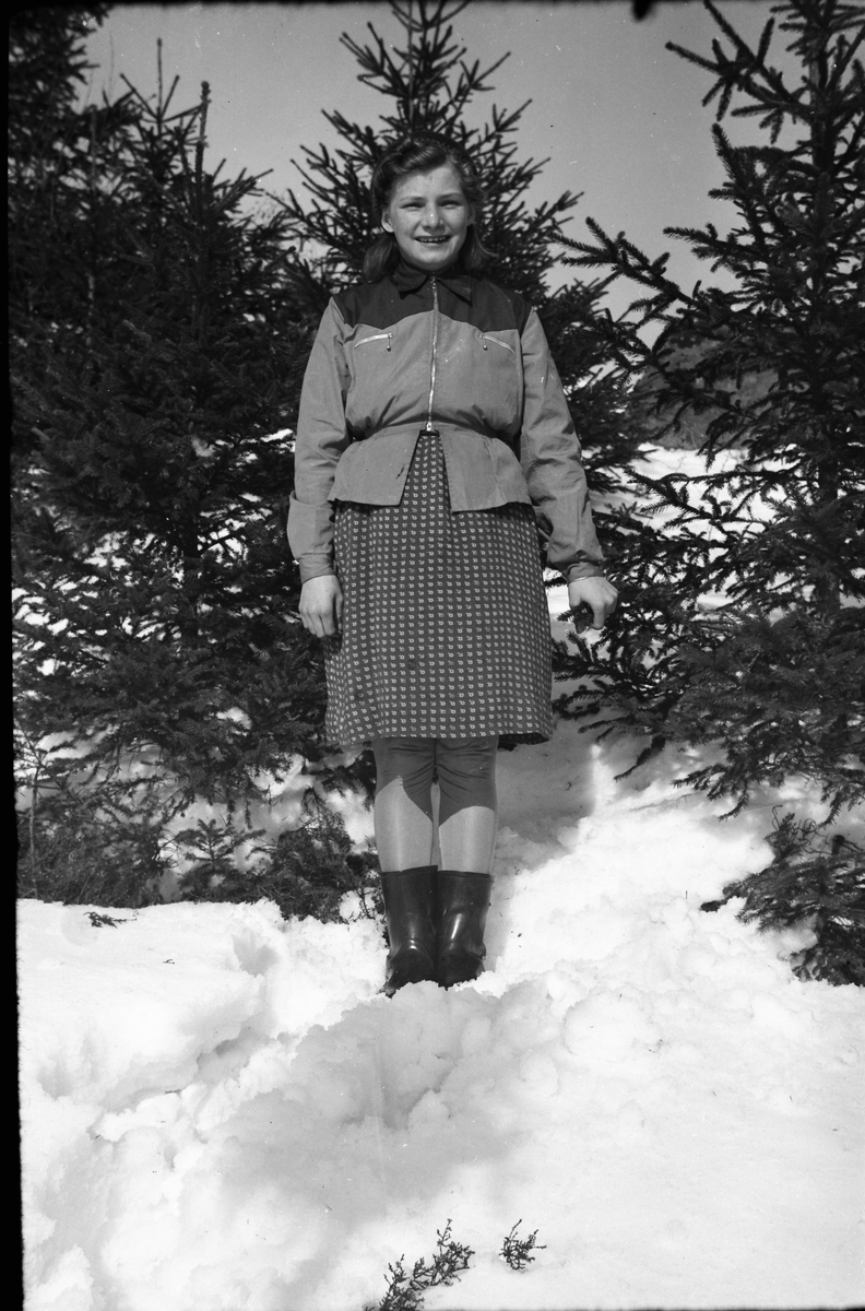 Ella Lundgren står vid smågranar i en snöbacke. Hon är klädd i midjekort jacka med blixtlås, kjol, strumpbyxor och gummistövlar.