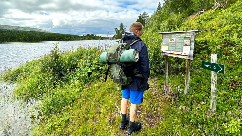 Bildet er utendørs. Man ser en mann i blå shorts, fjellsko og jakke som står å se på et informasjonskilt. Det er grønt bak skiltet og man ser vann ved siden av. På et lite skilt står det "post 16".