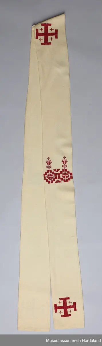 Et sett med messeklær bestående av svart prestekjole, hvit messeskjorte, kremgul og rød stola med kors, samt kleshenger av tre.