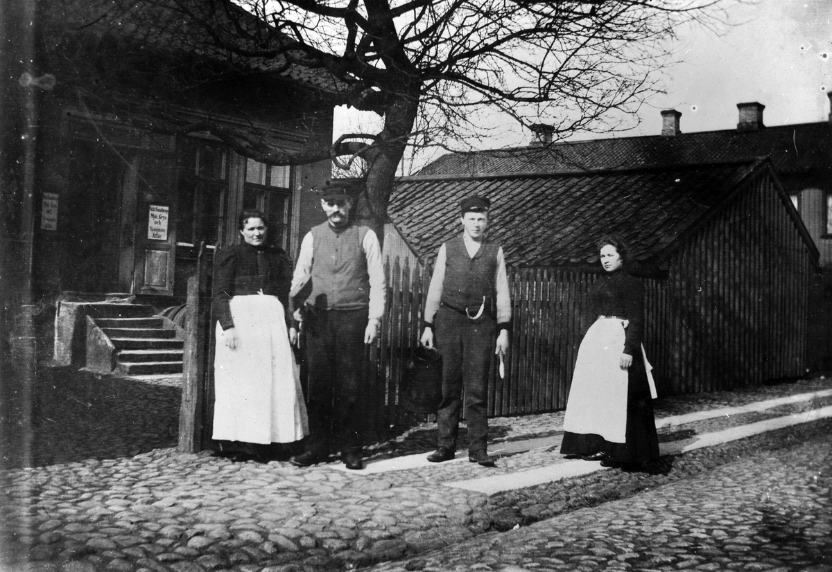 2 kvinnor, i ljust förkläde, och 2 män, i väst och keps, står på kullerstensgatan utanför Nils Sandbergs Mjöl Gryn och Spannmålsaffär på Nygatan i Kv Pärlan 6, tomt 74. Bredvid affären låg Brännvinsbolaget. I fastigheten bodde också barnmorskan Anna Bäckström. 
Mittemot (kv Jägaren 2, tomt 89) bodde enl husförhörslängderna handlaren Nils Sandberg. Han var handlande år 1859, 1865, 1876 (ej 1887) enl hfl. 

Fastigheten i kv Pärlan inrymde senare bl a mekanisk verkstad och sedan Westkustens Fisk. 
Fastigheten revs på 1980-talet och i den nyuppförda fastigheten finns bl a Westkustens Fisk och Föreningsbanken, samt bostäder.