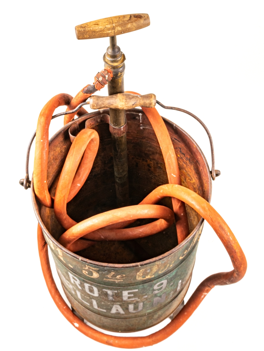 Brandspruta. Cylindriska behållare av järn, stående på två ben samt stöd. Pumphandtag i trä på ett rör av mässing och slang av gummi. Sprutmunstycke av mässing.