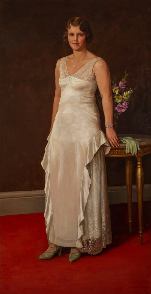 Helfigur porträtt av Milci Hesse i vit klänning lutad mot ett bord med vita handskar och en blomstervas