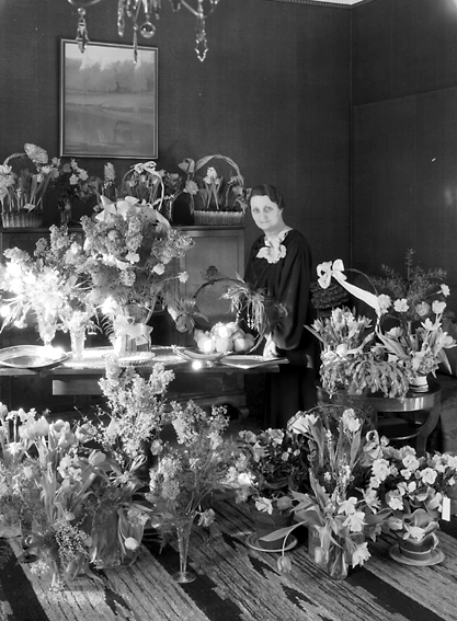Kvinna med fruktkorg omgiven av blommor.
Fotografens ant: Fröken Svea Andersson. 1936.