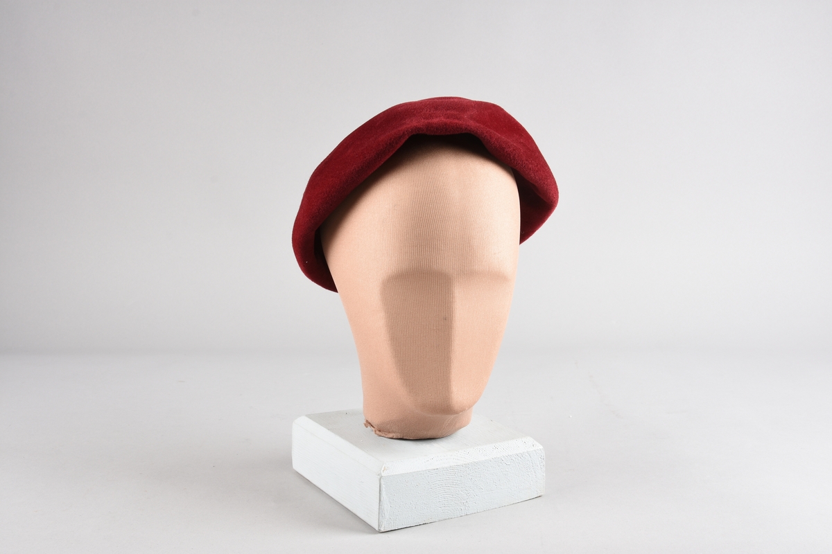 Rød, sirkulær hatt til dame, uten brem, med innbretett kant sydd rundt åpningen. Mulige små hull etter hattenål på den ene siden.