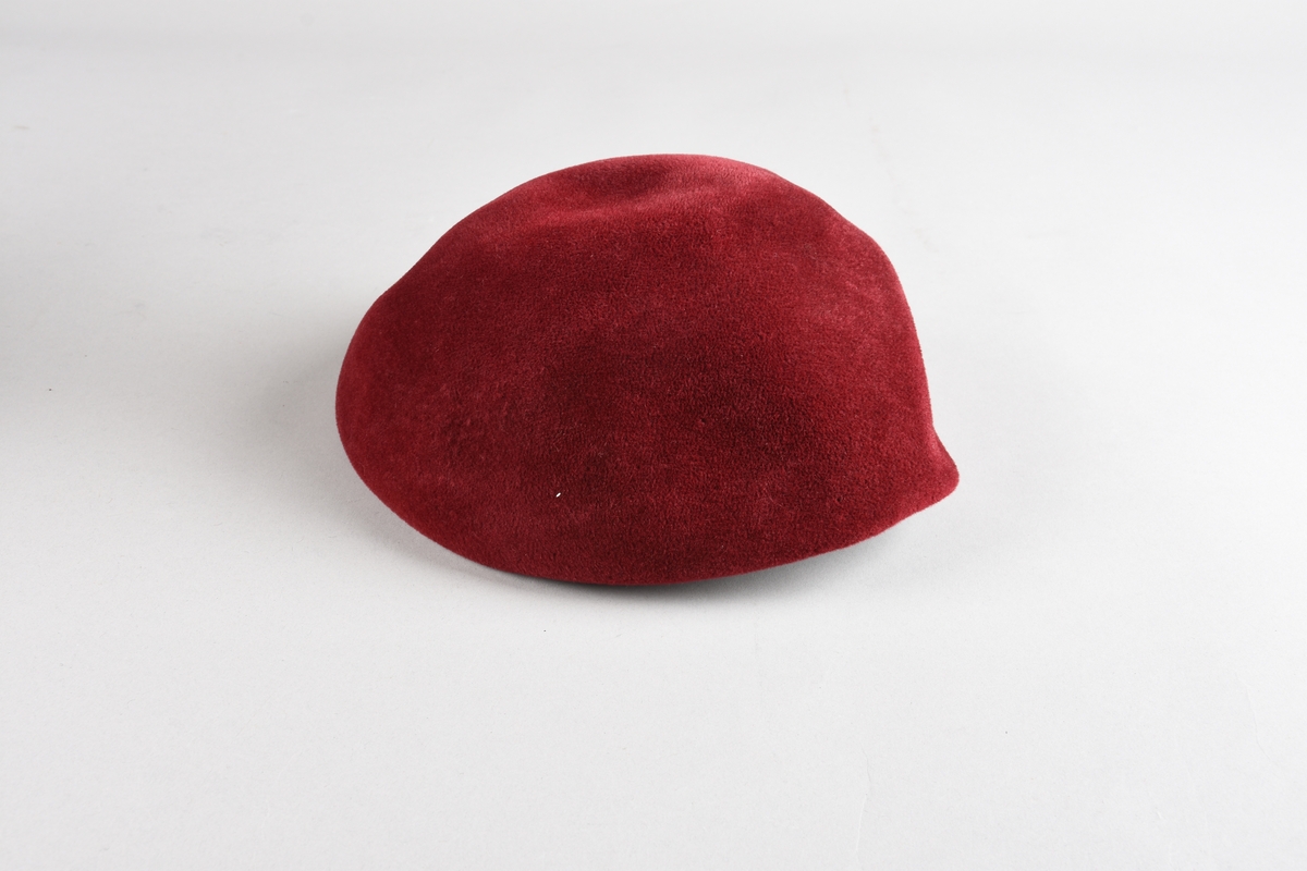 Rød, sirkulær hatt til dame, uten brem, med innbretett kant sydd rundt åpningen. Mulige små hull etter hattenål på den ene siden.