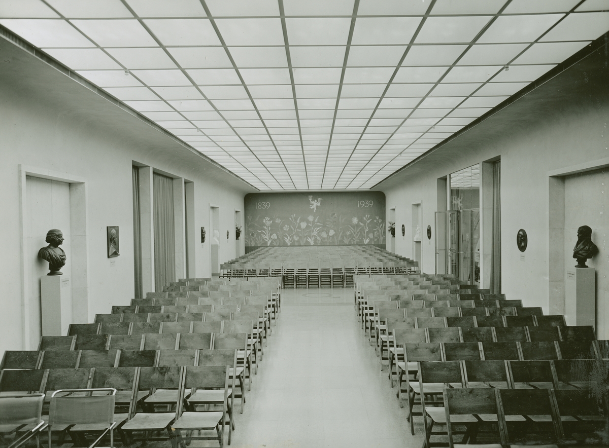 Tekniska museet, ca 1939. Sverigesalen, möblerad som föreläsningssal. Fonden dekormålad med en ängel med kandelabrar i händerna samt texten "1839 - 1939". Foto mot norr.