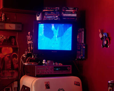 En skjerm troner i midten av et rom. Et kjøleskap og en videospiller. Et bilde av en mann vises opp-ned på skjermen..