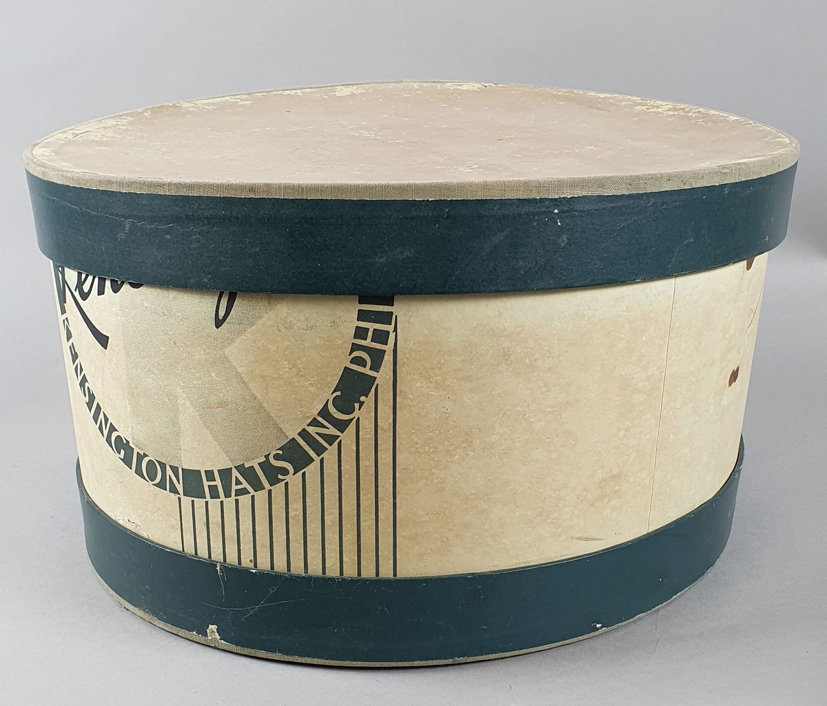 Sylinderformet hatteeske av kartong, med grønn dekor på utsiden. På innsiden er det en sylinderformet innsats med plate for en herrehatt.