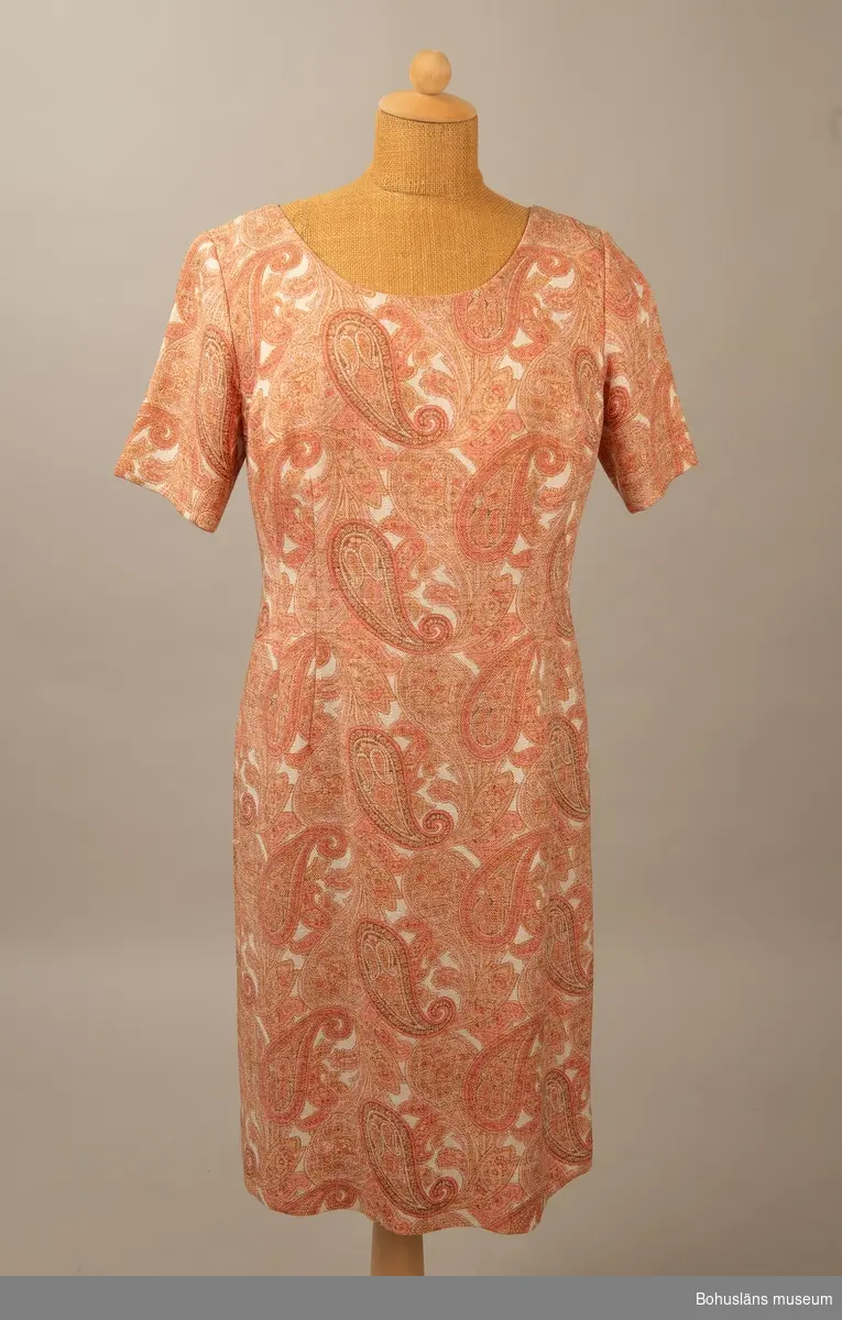Rak fodrad klänning i så kallat persiskt mönster. Milda rosa färger.
Insvängd i midjan och dragkedja bak.  Se bilaga.

Ärmlängd 10cm.