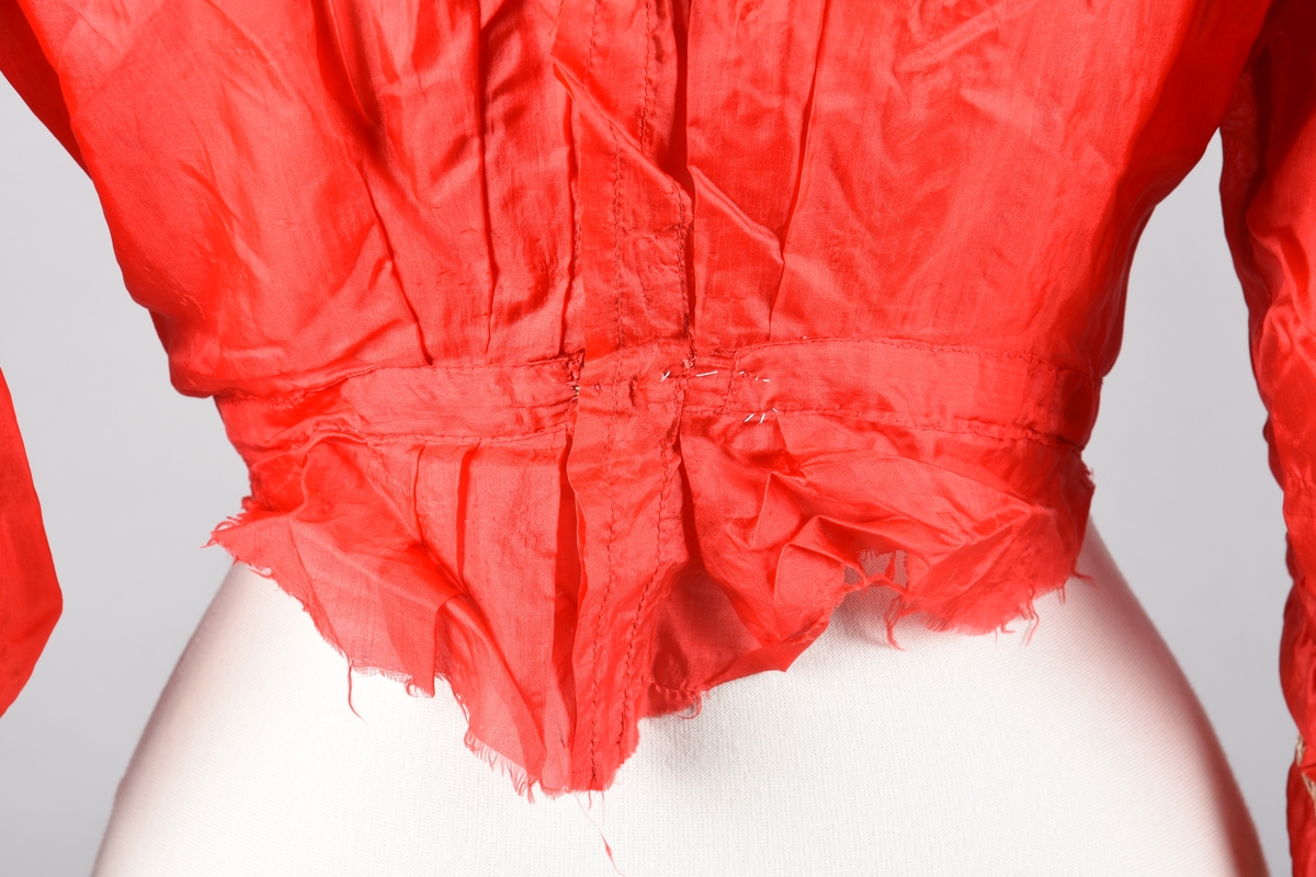 Bluse av tynn rød silke, åpen bak (hekter). Høy hals med spiler. Pyntet foran med kniplingsmotiv og hullsømsrader på armene og oventil.