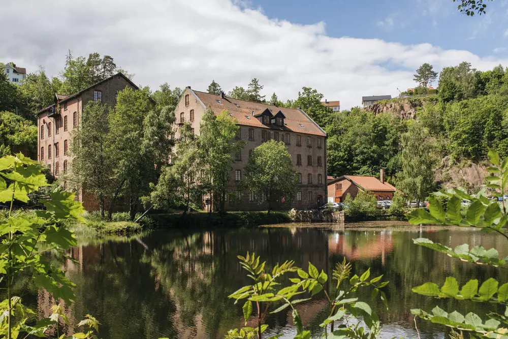 Berger museum i teglestein. Vi ser et vann foran bygget og mye grønt og trær rundt. Det er sol og sommer.