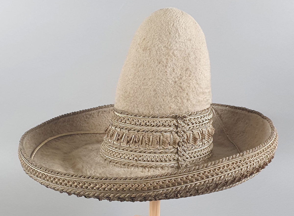 Beige (sauehvit) meksikansk hatt av børstet ull. Hatten har høy smal pull og bred brem som vender oppover. Flettede brede bånd av strå med sølvtråd rundt pullen på yttersiden og bremmen på undersiden. Smalt kantbånd ytterst på bremmen har nesten revnet helt av.