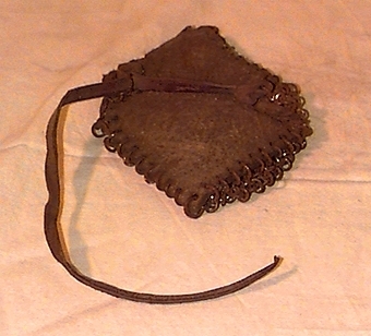 Fyrkantigt ylletyg som är beklätt med ett nät av cirkelformade metallringar på ena sidan, och har en läderrem på andra sidan.