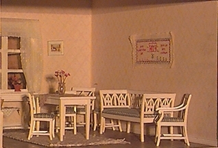 Väggar med tryckta tapeter. Ett fönster med spröjsade rutor, gardiner i form av kappa och längder. Dörr med tre speglar. Golvet av plywood, taket målat. Sittmöbel, vitmålad med blårutig klädsel bestående av soffa (jfr 34 081, ej lyra) med figursågade rygg- och armstöd, två karmstolar samt fyra stolar med samma figursågning som soffan, bord 
(jfr 34 069) med duk och vas med blommor. Gungstol med matta i. Under fönstret ett bord med låda. Byrå med tre lådor, vitmålad med guldfärgade beslag, på byrån står två ljusstakar med ljus samt ett keramikfat. Ett litet slagbord med en duk på, därpå en korg med rött garnnystan i samt en sax. Två krukväxter i fönstret. Tre tavlor varav en Carl Larsson och en Prins Eugen. Ovanför byrån en oval spegel i guldram. Broderad bonad ovanför soffan. På golvet en vävd matta i vitt och blått.
Belysning i form av en glödlampa vid framkanten insatt vid senare datum då skåpet placerats i ett montage med skåp nummer 34 048-34 054.
