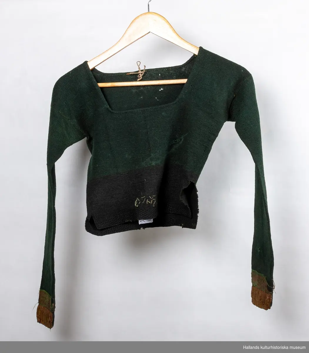 Grön stickad tröja i ull med svart nederkant. Sidenband på ärmlinningarna. Längd från axeln 40 cm. Längd på ärmen 50 cm. Midjevidd 66 cm.