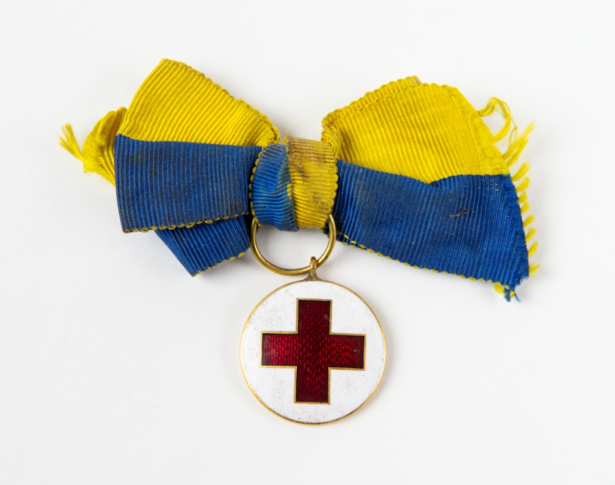 Medalj, Röda Korset med blågul rosett. Medaljen fästes på baksidan med en säkerhetsnål.
