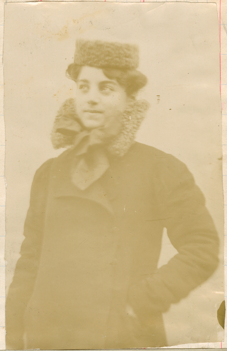 Ung kvinne fotografert utendørs. Elegant antrukket i en kåpe med matchende hals og lue i ull. Sløyfe i halsen.