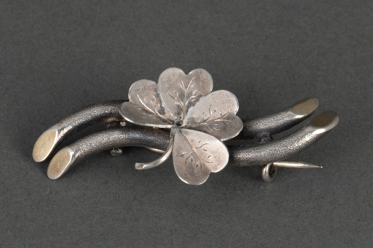 Nål/ brosje i sølv. Et firkløverblad er montert på to grenlignende rør. Gravyr på bladene. På smykkets underside er det festet en nål.