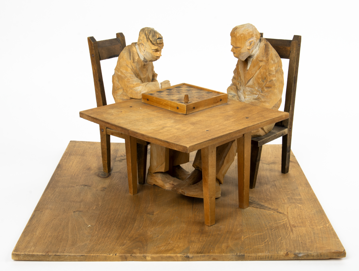 Skulptur av trä av föreställande två schackspelare sittande på var sin stol vid ett bord.