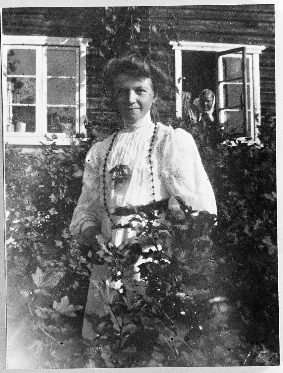 Marie (fremst, innimellom buskene i hagen) og Andrine Skinderviken (bak, i vinduet). Bildet er tatt 15/9-1907.