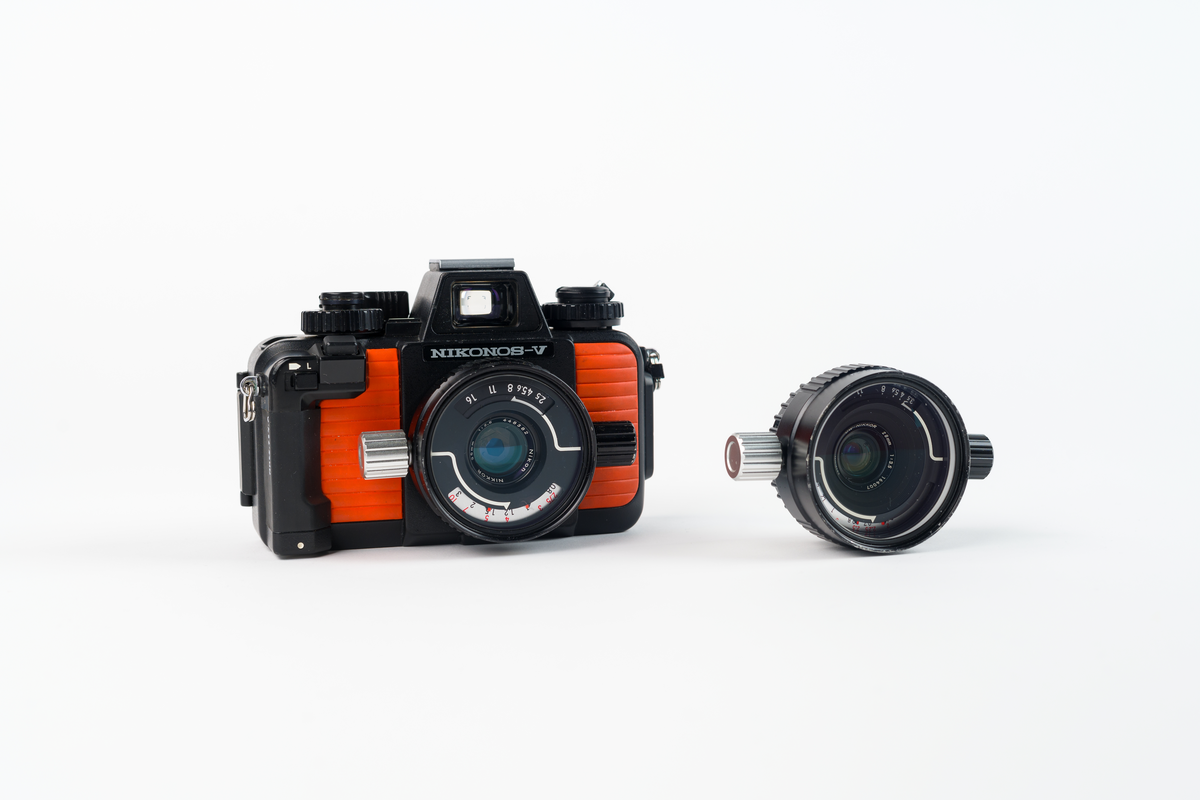 Undervattenskamera Nikonos V. Svart kamera med detaljer i orange. Extra objektiv bredvid. Kameran märkt i vitt: "Nikonos-V"