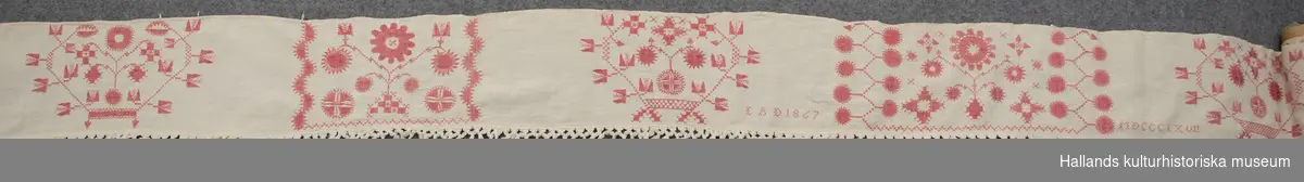 Hängkläde i linne. Broderat i geometriska mönster, rött på vit botten med påsydd knuten, vit frans. Märkning: "EAD", "1867", "MDCCCLXVII". Frans +:70 mm.