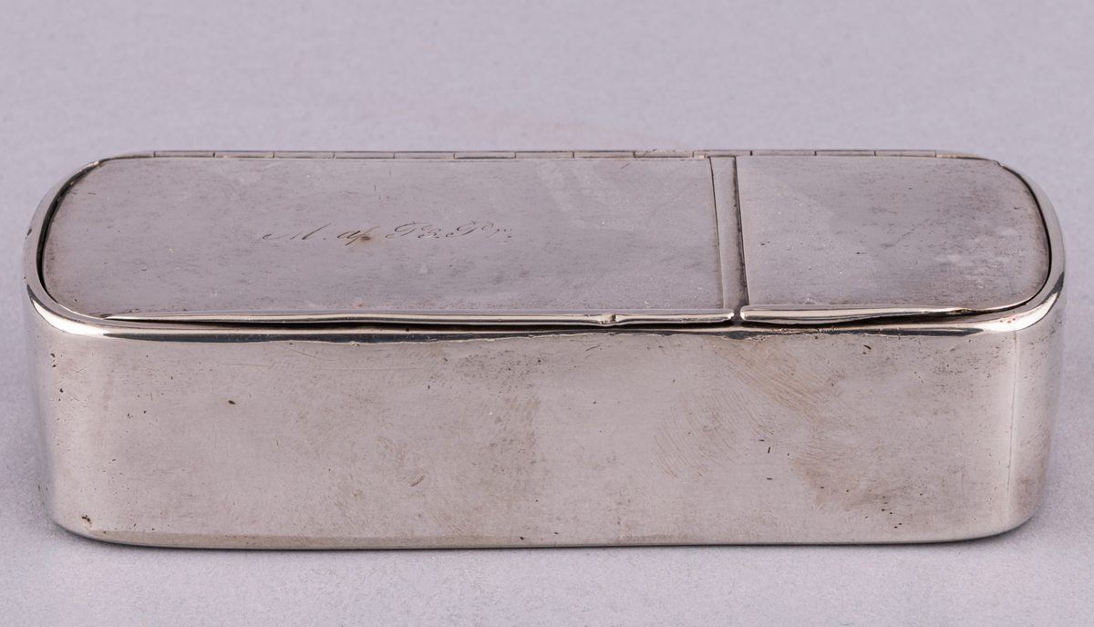 Snusdosa i silver, indelad i två fack och försedd med lock.Rektangulär med rundade hörn. På ett av locken ingraverat M af P3 Pr. Tillverkad i Gävle. Stämplarna otydliga.
