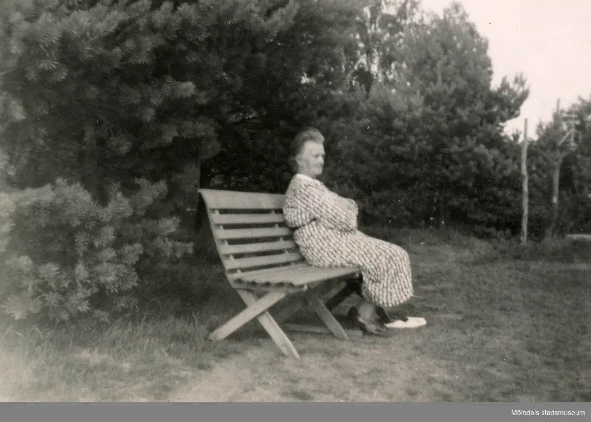 Nora Krantz (1879 - 1955) sitter på en bänk i trädgården utanför hemmet (Stretereds personalbostad på Tulebovägen 23-27 i Stretered). Nora var gift med skomakaren Carl Krantz som arbetade på Stretereds skolhem. Nora och barnen flyttade till det då nybyggda huset i Stretered 1921. Carl flyttade dit tidigare, först inhyst i Sporred.