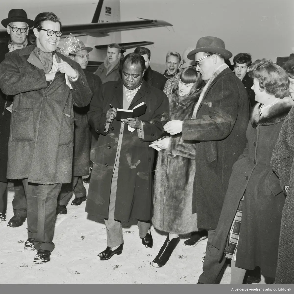 Louis Armstrong ankommer Oslo via Fornebu flyplass 2 februar 1959. Ønskes velkommen og intervuet av blant andre Haagen Ringnes (th) og Tor von Krogh. Ytterst til venstre: impresario Per Gottschalk