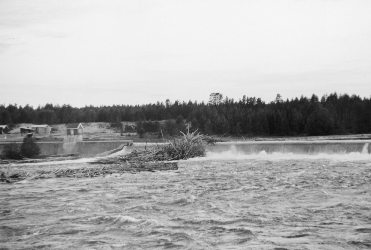 Tømmervaser ved kraftverksdammen ved Skjefstadfossen i Heradsbygda i Elverum i Hedmark. Fotografiet ble tatt i 1938, fra østre elvebredd langs den buete overløpsdammen. Her ser vi, til høyre i bildet, den vestre delen av den 312 meter lange betongmuren med avrundet krone som skulle stuve opp elvevannet og på den måten magasinere over 600 millioner kubikkmeter vann. Fløtingstømmeret på motstrøms side av dammen ble forsøkt ledet mot den østre elvebredden, for der var elveløpet djupest, noe som reduserte faren for at stokkene skulle sette seg fast og stuves sammen i hauger. Målet var at tømmeret kunne flyte uhindret videre nedover med strømmen. Da dette fotografiet ble tatt hadde det bygd seg opp en haug av tømmerstokker som sprikte i mange retninger under den vestre delen av overløpsdammen. Her hadde det lagt seg en haug under dammen, og etter hvert som mer kom ovenfra bygde haugen seg opp over dammen. I 1938 var det innmeldt 4 436 159 tømmerstokker til fløting fra den delen av Glommavassdraget som lå ovenfor Skjefstadfossen.