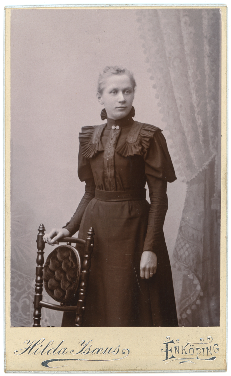 Ateljéfoto av en ung dam stående vid en stol. Baksidan av visitkortet är blankt.