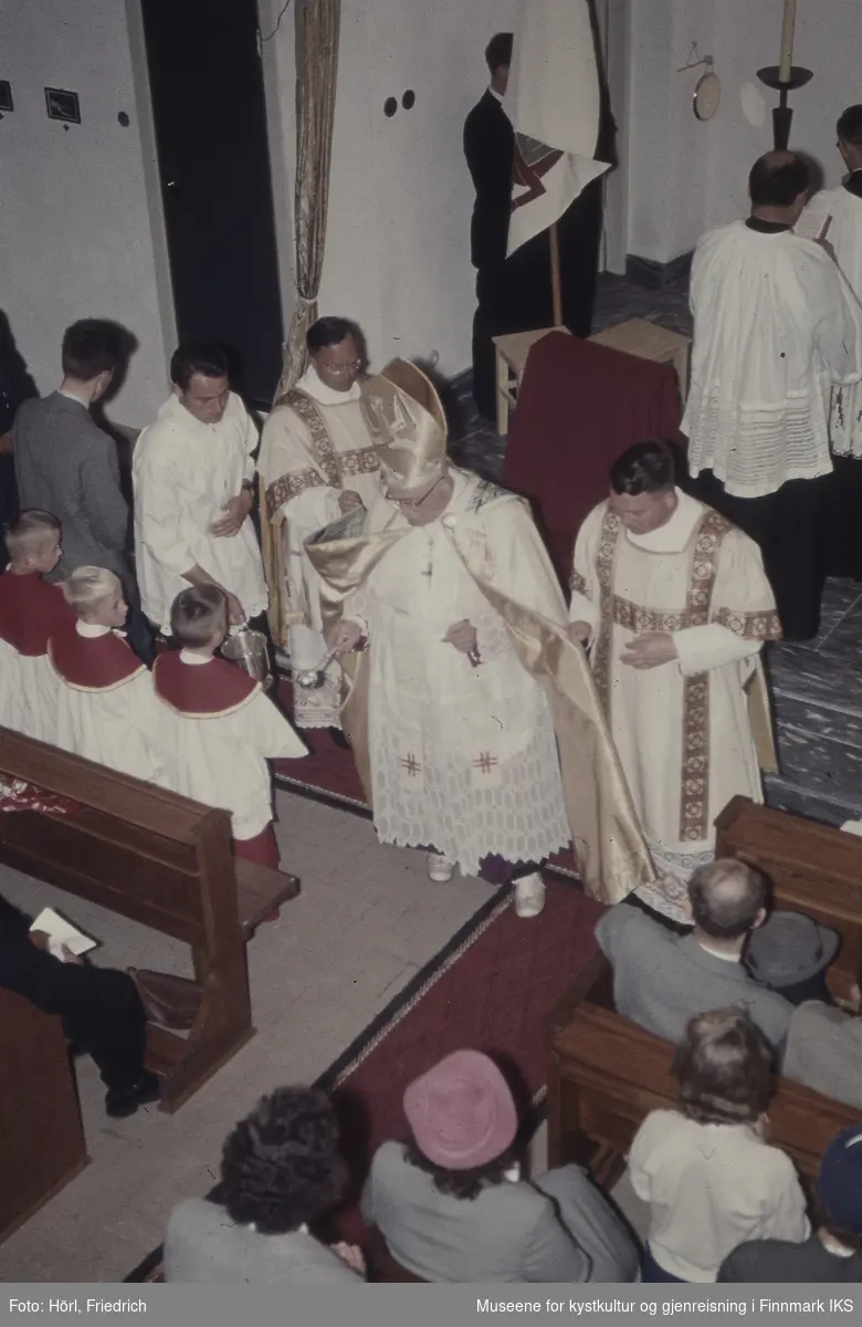 Bildet er tatt fra galleriet under innvielsesgudstjeneste av Den katolske kirke St. Mikael i Hammerfest den 03.08.1958. Det vises Biskop Wember som holder en øse med vievann i hånden og hans følge, kirketjenere og en del av menigheten.