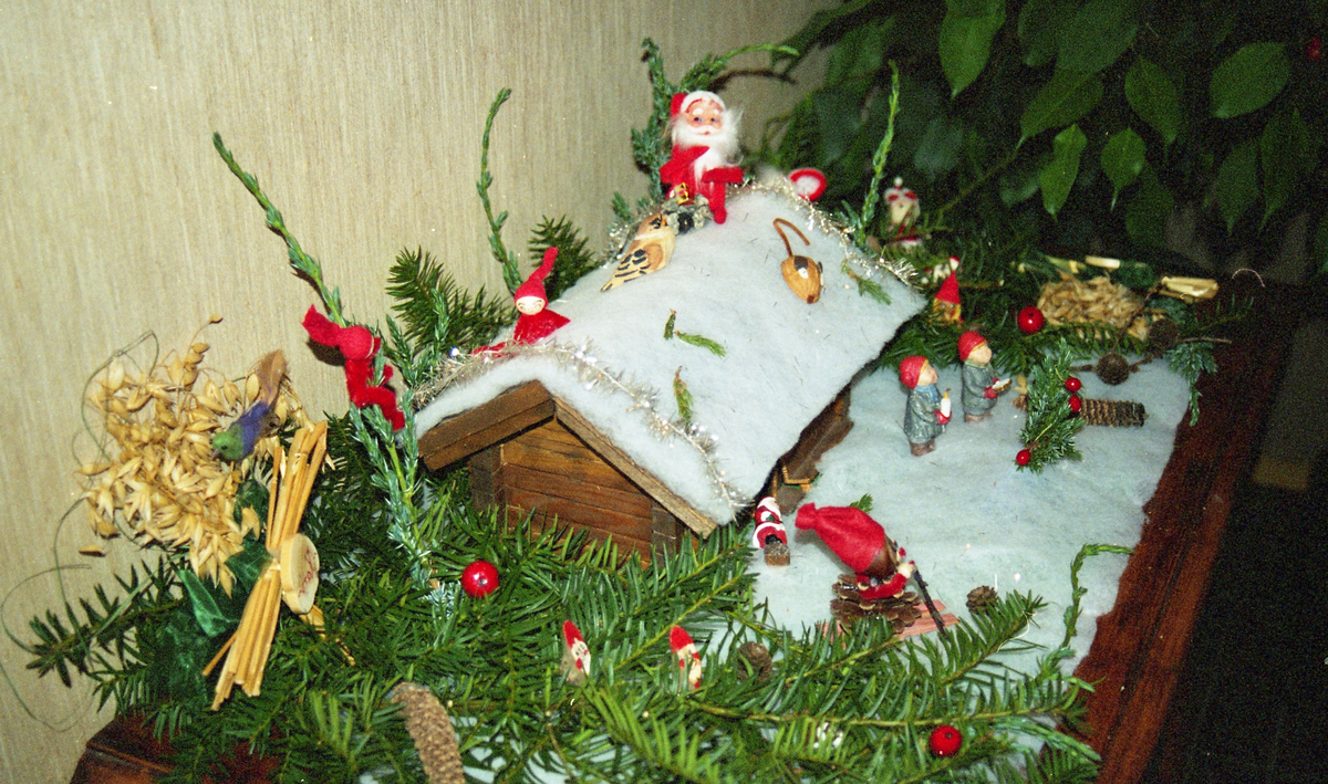 Eit lite julepynt-tablå: eit lafta hus med trær av granbar rundt. "Snø" av bomull på taket og bakken. Små figurar, nissar og mus plassert rundt omkring.
