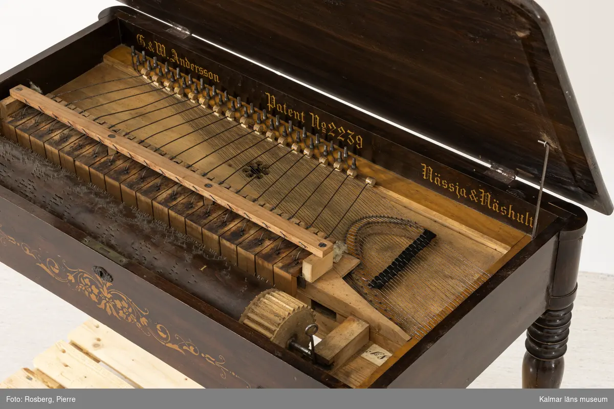 KLM 25434. Pianoharpa, vevpiano. Rektangulärt brunbetsat och lackerat bord med svarvade ben. På sargen schablonmåleri i guldfärg. Bordsskivan kan öppas, inuti en inbyggd melodivals som drivs med en löstagbar vev. Valsen kan justeras i sidled för att spela olika melodier. Inuti text i guld: G. & W. Andersson Patent No 2239 Nässja & Näshult. Intill valsen en papperslapp med text: No 89.