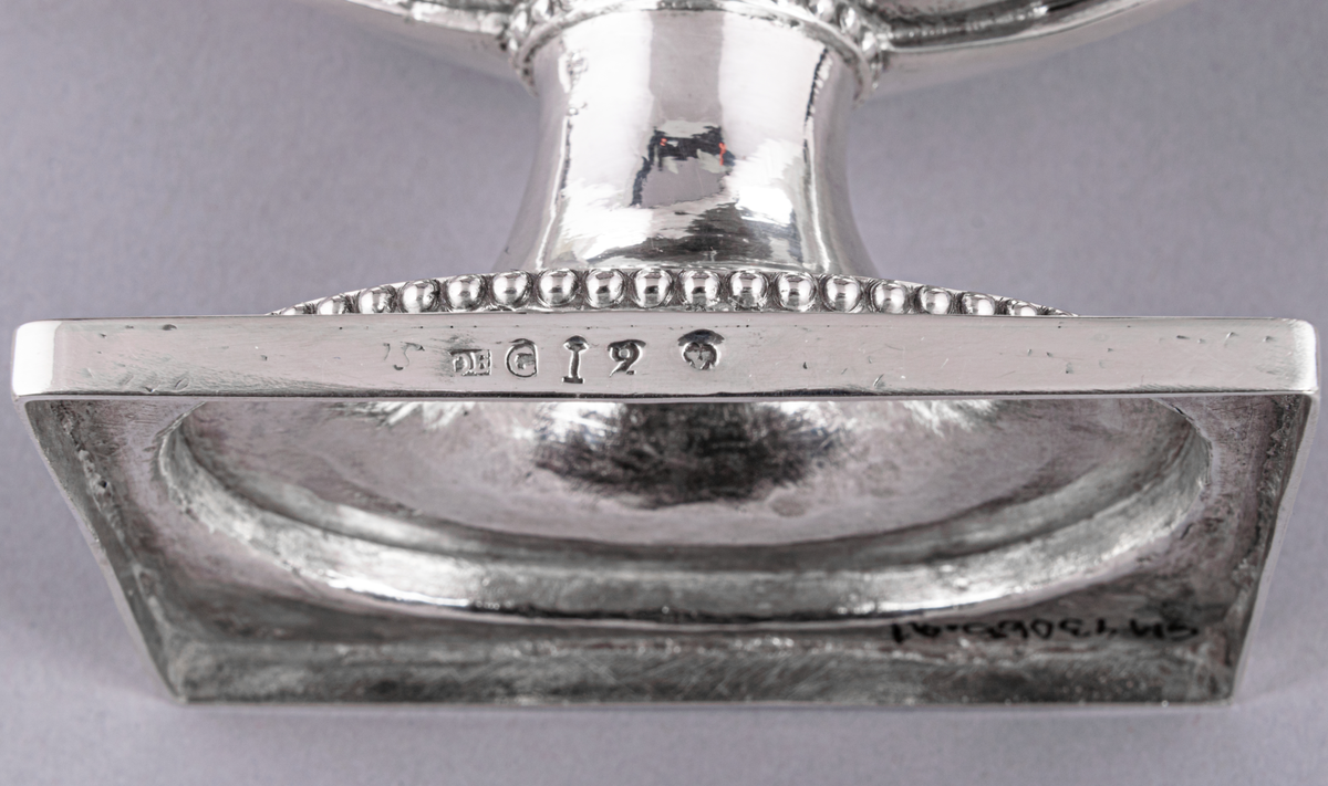 En av ett par sengustavianska sockerskålar i silver stämplade Daniel Elfbom, Gävle 1791. Oval skål på rektangulär fot. Löst lock med knopp och två utvändiga uppsvängda hänklar med pärlstav. Pålödd medaljong med Gävle stads vapen (G). Stämpar på fotens kant: DE G I 2 och tre kronor.
