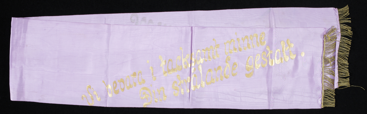 Violett sidenband med guldtext lydande; "Se vi bevarar i tacksamhet minne din strålande gestalt/Kvinnorna av Värend".

Inskrivet i huvudkatalog 1983.