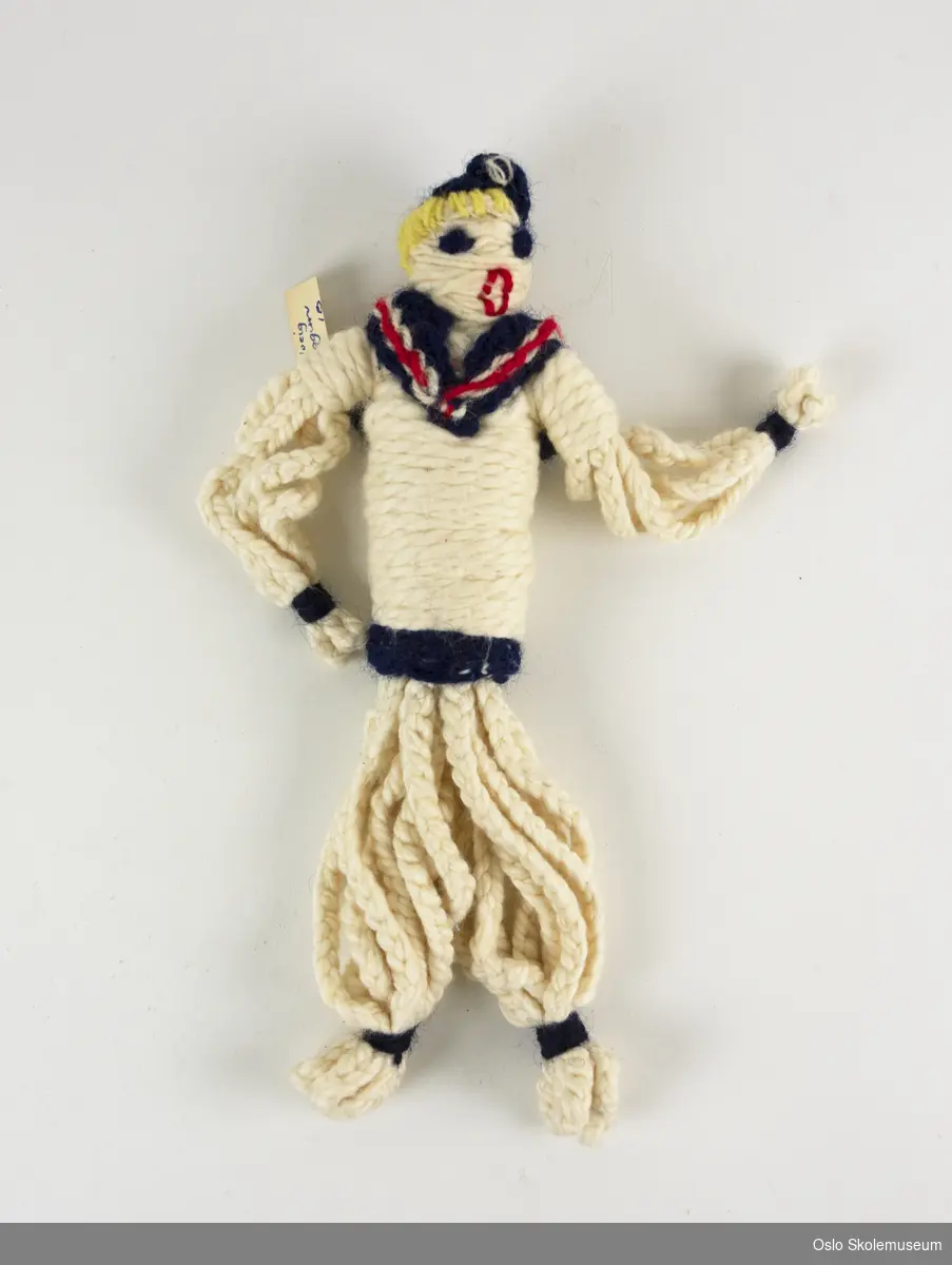 Dukke laget av surret og flettet tråd. Dukken er en sjømann med hvite klær med blå og røde detaljer. Den har også gult hår og blå hatt.