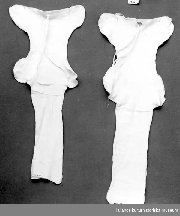 Två stycken barnblöjor av vit bomullstrikå. Konfektion. Två hopsydda lager, blöjformad. På ena änden ett påsytt rektangulärt stycke som viks över blöjan och fungerar som stjärtlapp. Knyts fast med bomullsband.