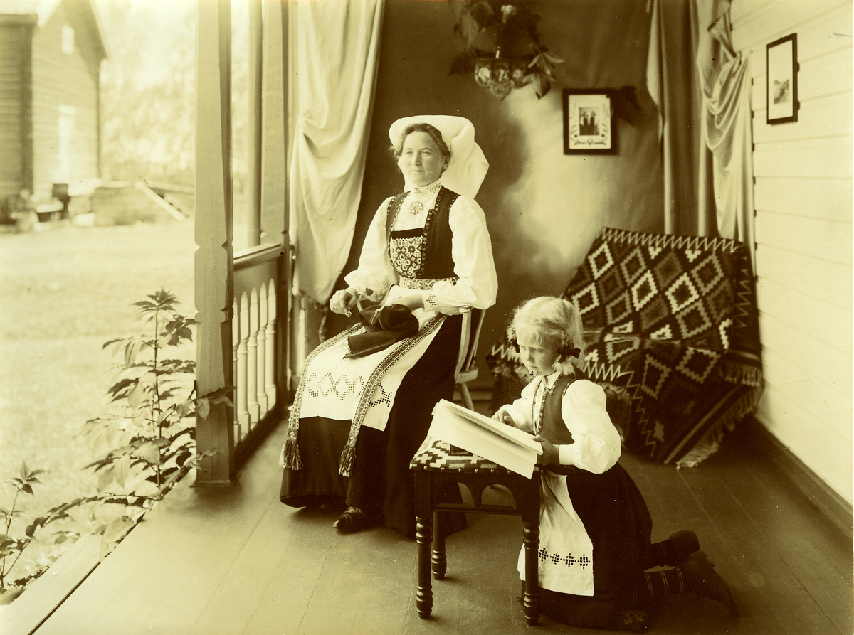 Mor og datter i Hardangerbunad. Mora broderer og dattera leser i ei bok. Hodeplagget tyder på at dette er bunader fra Sørfjorden.
