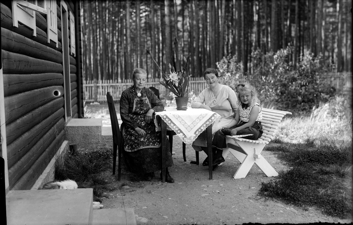 Gruppebilde.Ved et bord i hagen sitter mor til Alma,Alma og Gard Langslet. Fin duk på bordet.
