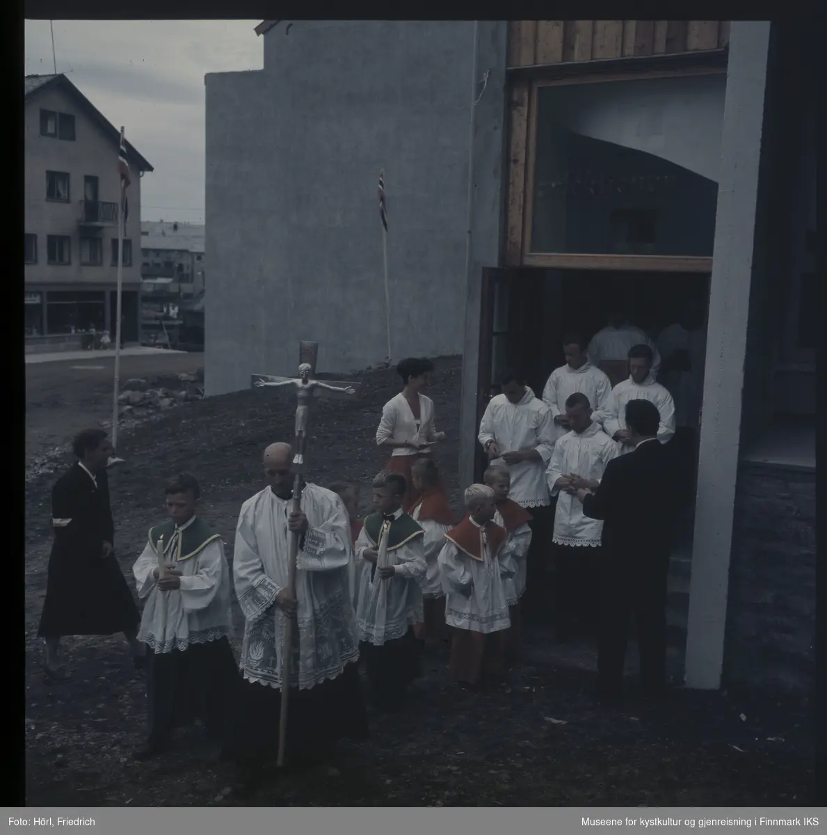 Presten bærer prosesjonskorset og leder an toget ut av Den katolske kirke St. Mikael i Hammerfest i 1958. Ministrantene går bak ham og to av dem holder stearinlys.