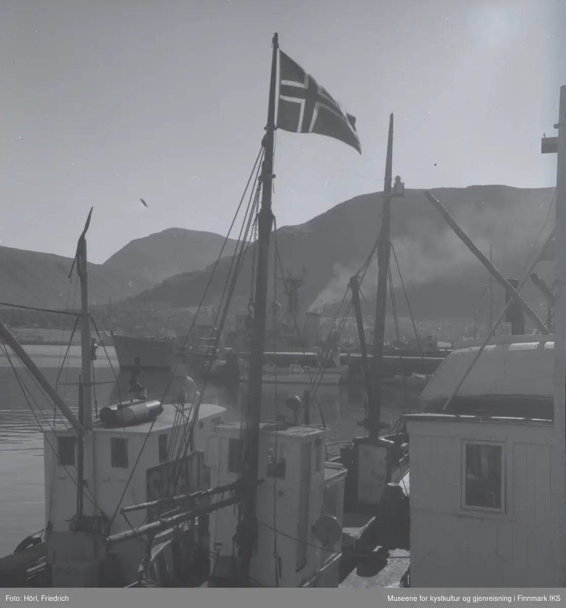 Båter av ulike typer ligger til kais i Tromsø. I forgrunnen ser man en fiskebåt med det norske flagget heist i masta. I bakgrunnen ser man Fløyafjellet.