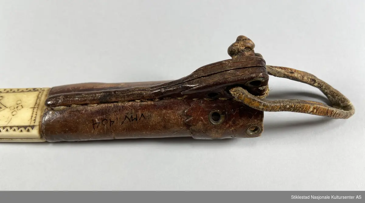 Knivslire laget av reinshorn og lær. Klinket med kobbertråd. Messingmaljer øverst på slirelæret som dekor. Inspirert av samisk dekor. Knivsliret er merket med I.J. 1934