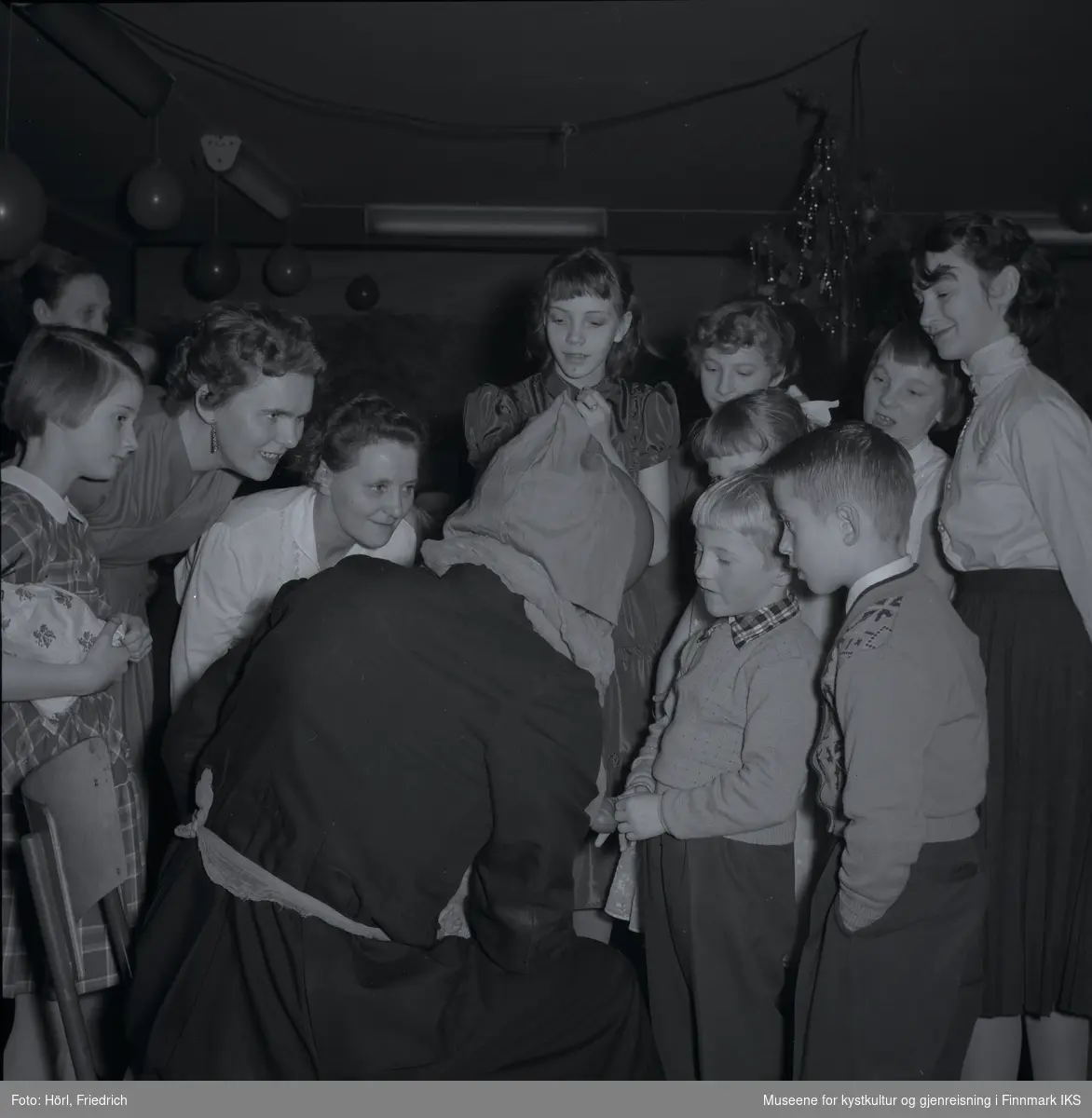 Julenissen besøker juletrefesten i den katolske menigheten i Hammerfest i 1957. Nærmest julenissen står det to gutter. En av dem ser ut som om han sier opp en dikt eller noe lignende for å få en gave. Dette er Terje Trondsen. De voksne kvinner ser på og er tydelig glade, spente og opprømte. Jenta til venstre i bildet holder sin presang i armen allerede. I bakgrunnen ser man juletreet og luftballonger.
Noen av de avbildete voksne er Ellionore Eriksen og Gerd Trondsen.