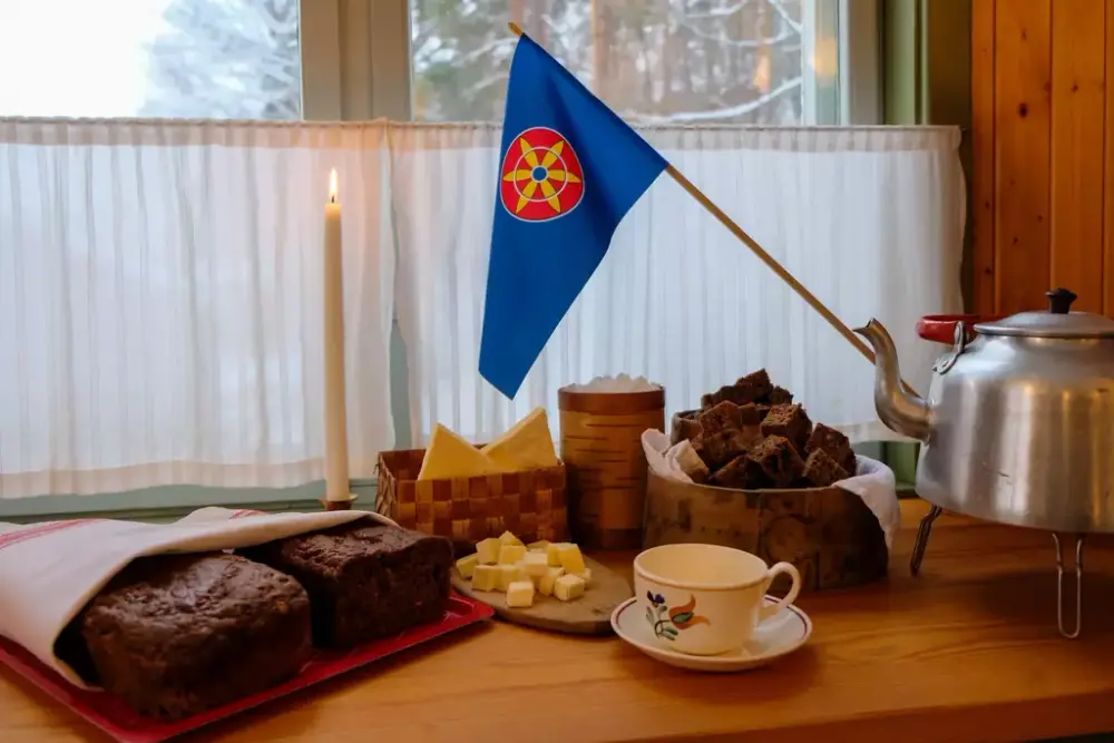 Et bord dekket med tradisjonell kvensk mat, inkludert brød, kaffeost, kaffekanne og en kopp. Det kvenske flagget og et tent stearinlys skaper en varm og koselig stemning. 