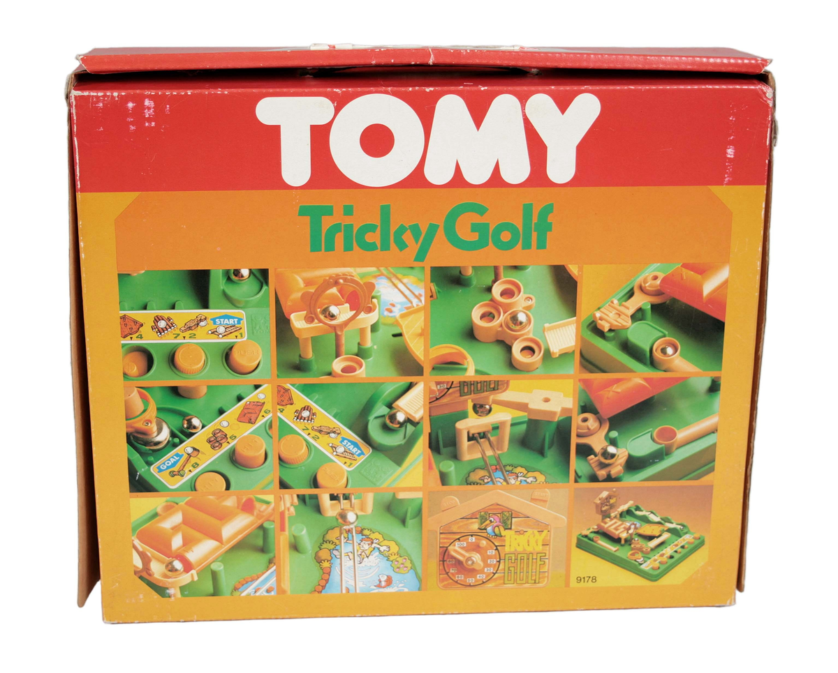 Minigolfspel i plast. Grön spelplan med detaljer i beige, orange och brunt. Metallkula. Instruktionsbilder i färg. Tidur. I originalkartong av papp märkt "TOMY Tricky Golf. Made in Singapore".