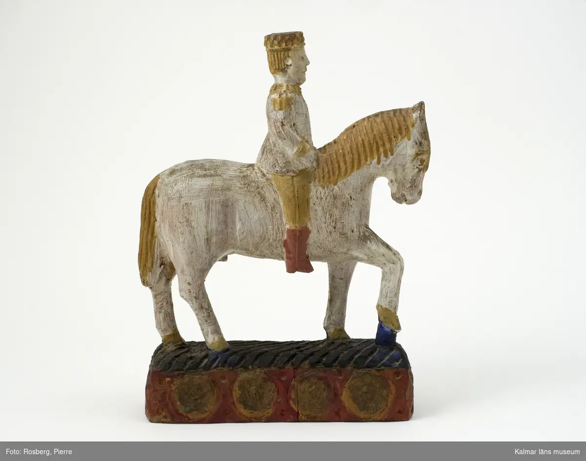 KLM 12928. Skulptur, träskulptur. Skulptur i form av en manlig ryttare på en häst. Ryttaren och hästen bemålad i vitt, gult och rött. Hästen står på en sockel bemålad i rött, gult och blått. Titel: Faraos ryttare.