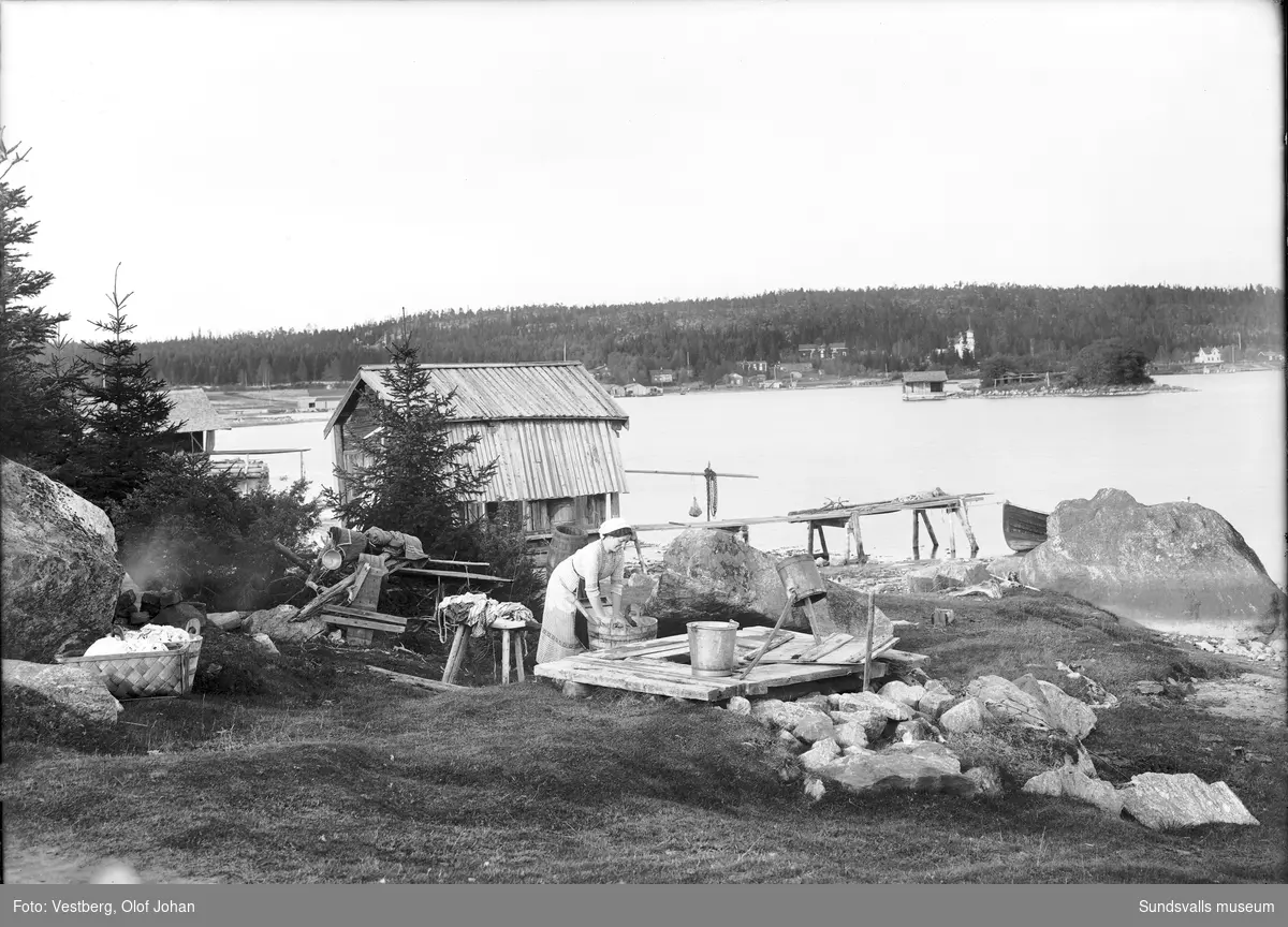 En kvinna står och tvättar kläder invid en sjöbod i Bodviken. Längre bort ön Lillrukan och längre bort syns Juniskärs herrgård, senare pensionat. På bild två samma sjöbod ur en annan vinkel.