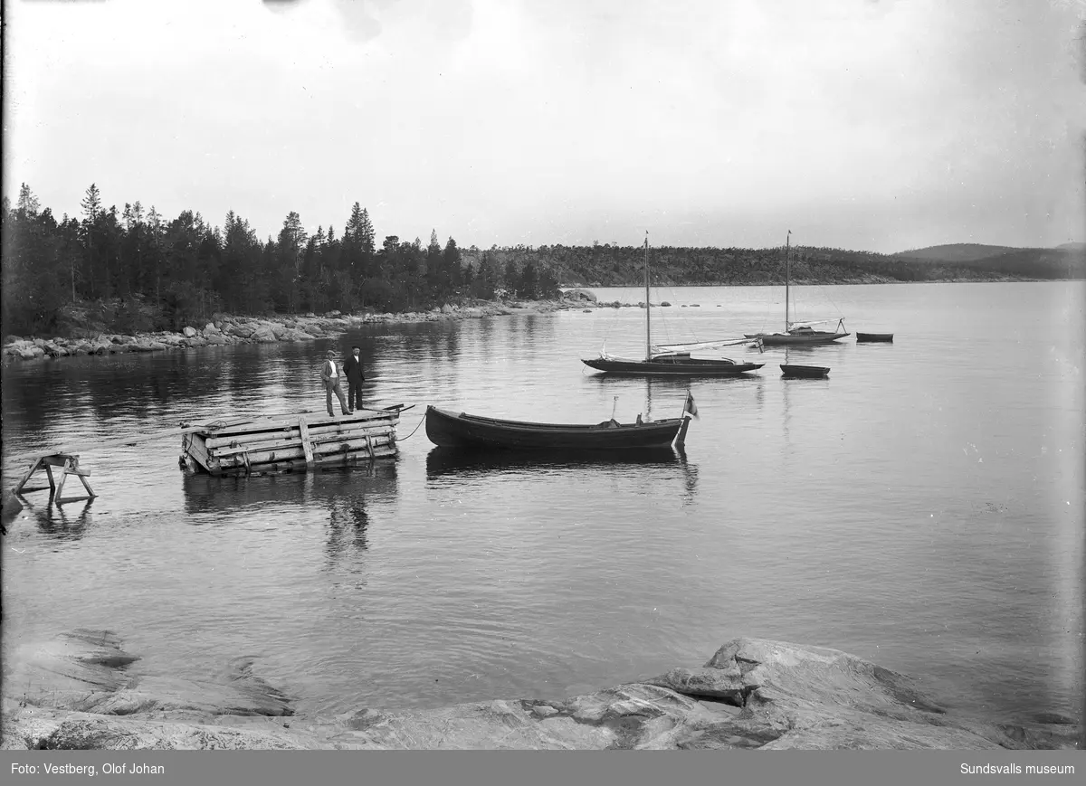 Vy från en havsvik i Juniskärs skärgård, troligen nordvästra sidan av ön Skigan. Tre båtar ligger för ankar och två män står på en brygga.