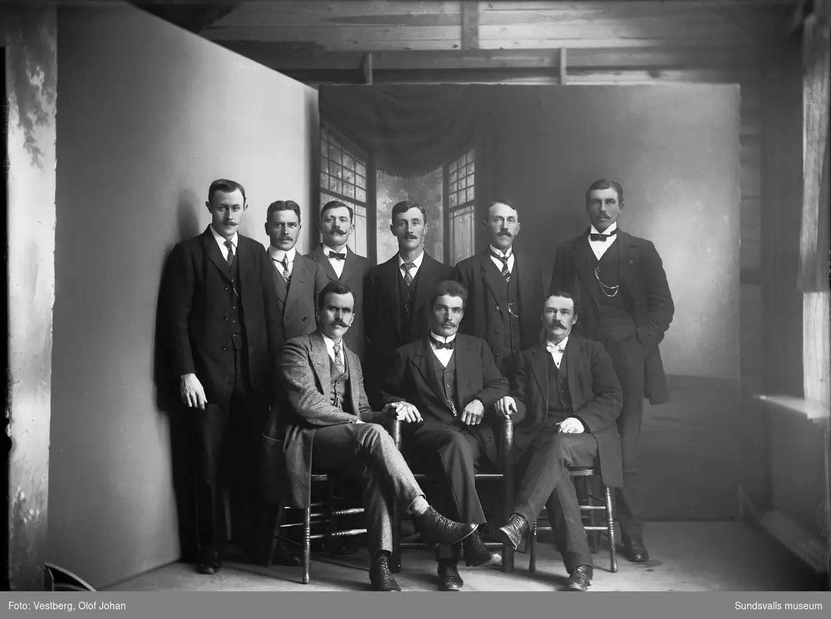 Ateljéfoto med en grupp på nio män i mörka kostymer.